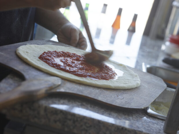 Pizza chef spreading tomato sauce around pizza dough on a peel in a restaurant
