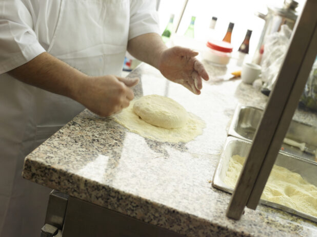 Pizza chef in a white apron with a ball of white dough in cornmeal about to make a pizza in a restaurant kitchen
