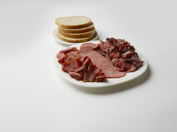 Three types of sliced deli meat on a plate with rye bread on a white background