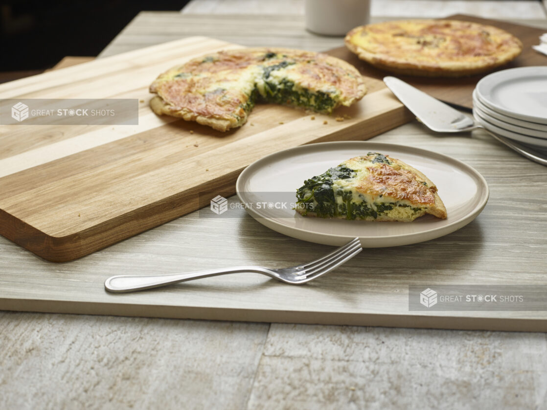 Slice of spinach and cheese quiche on a plate with two quiches in the background on wooden boards