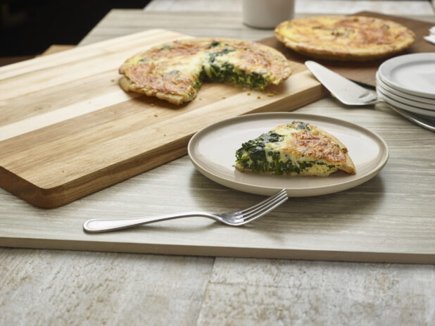 Slice of spinach and cheese quiche on a plate with two quiches in the background on wooden boards