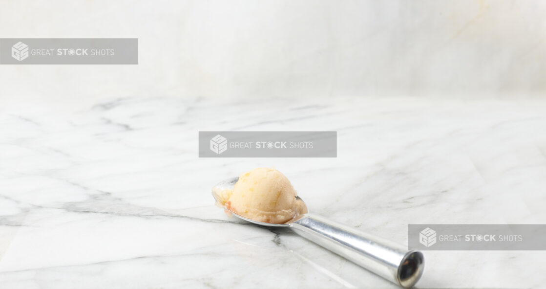 Scoop of peach gelato on a silver scoop with a white marble background