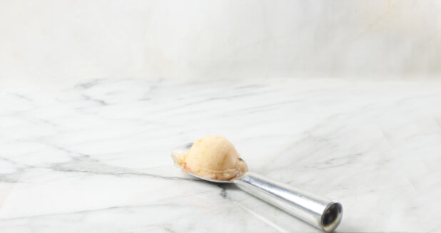 Scoop of peach gelato on a silver scoop with a white marble background