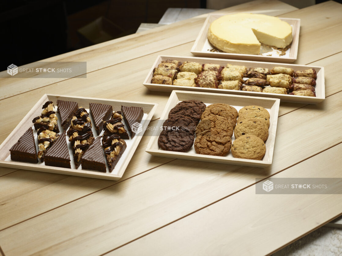 Assortment of cookies, squares, pastries and a plain cheesecake in wooden trays on a wooden background