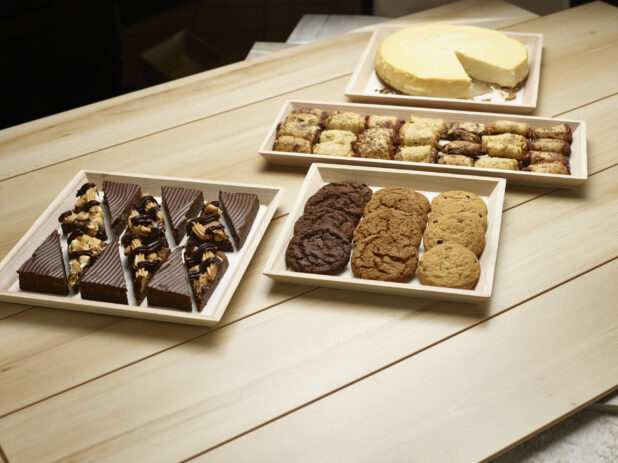 Assortment of cookies, squares, pastries and a plain cheesecake in wooden trays on a wooden background