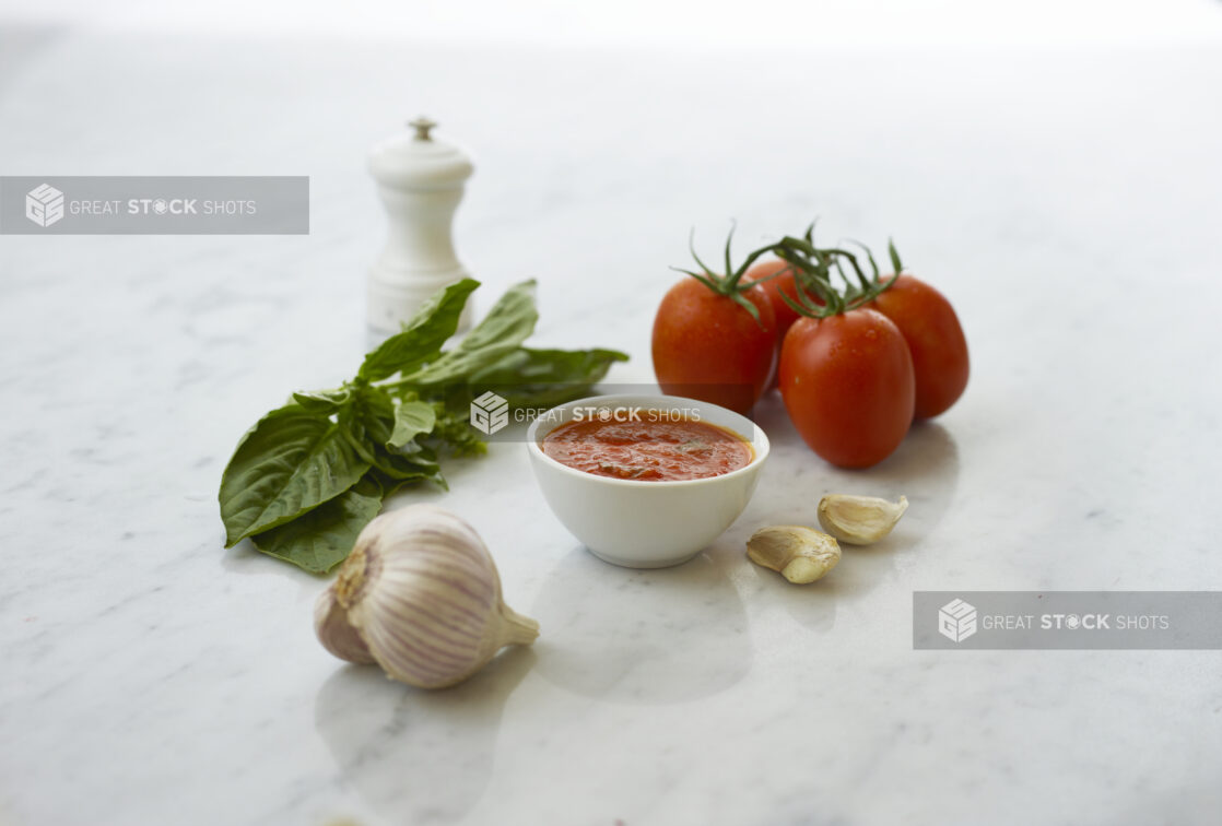 Small white bowl of tomato sauce surrounded by ingredients on white marble