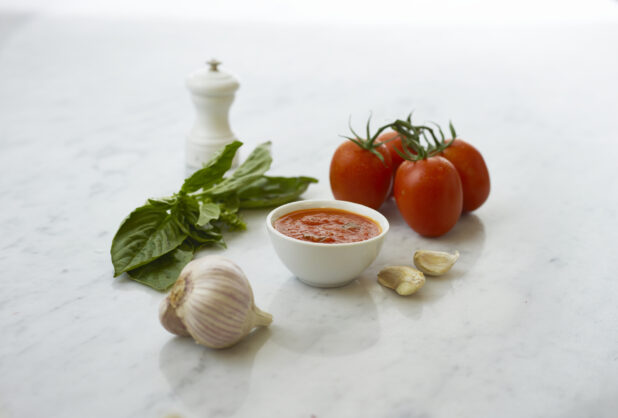 Small white bowl of tomato sauce surrounded by ingredients on white marble