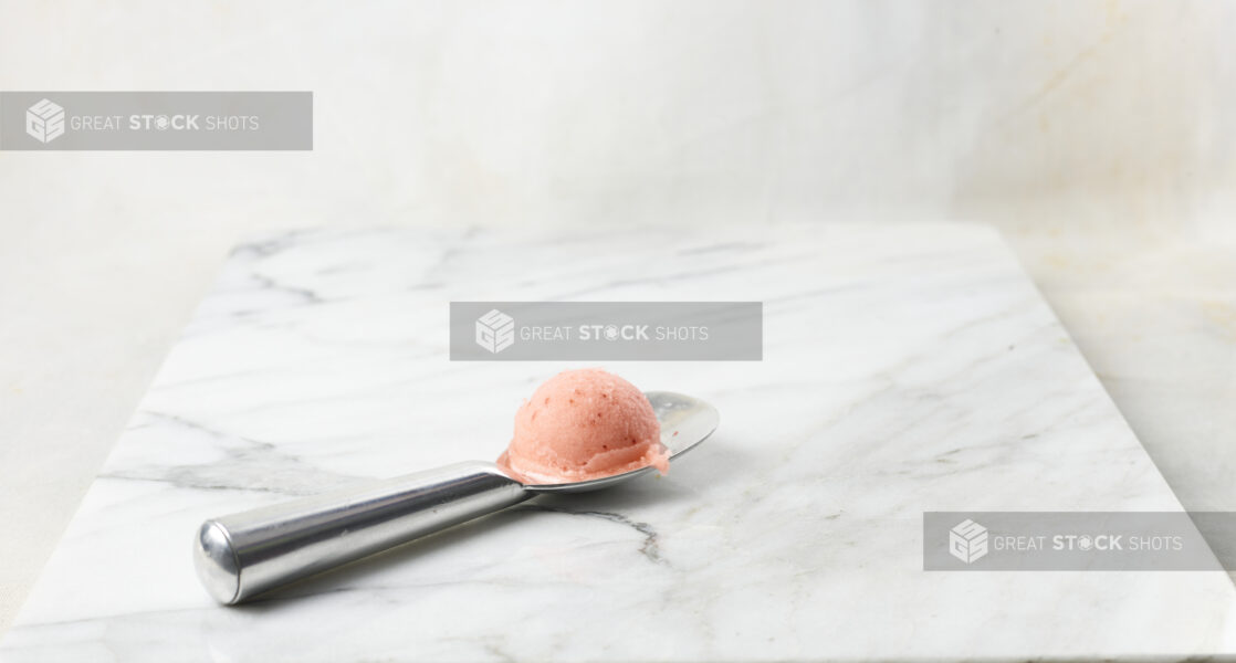 Scoop of strawberry gelato on a silver scoop with a white marble background