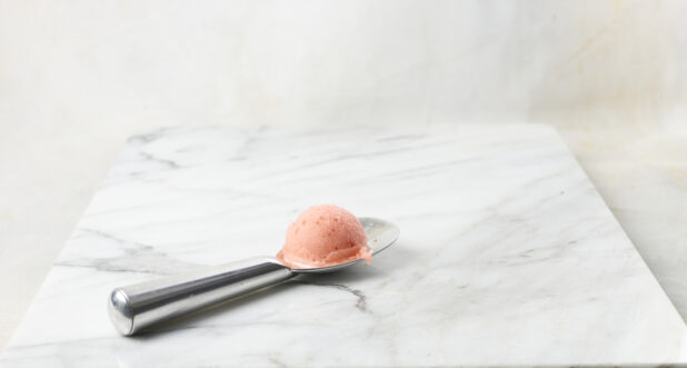 Scoop of strawberry gelato on a silver scoop with a white marble background