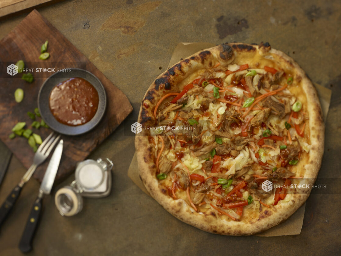 Whole medium pulled pork pizza with red peppers, cabbage, onion and shredded carrot on parchment paper with barbecue dipping sauce on the side, overhead view