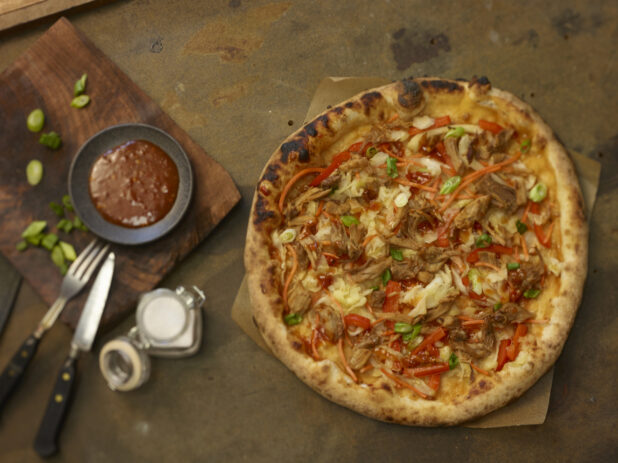 Whole medium pulled pork pizza with red peppers, cabbage, onion and shredded carrot on parchment paper with barbecue dipping sauce on the side, overhead view