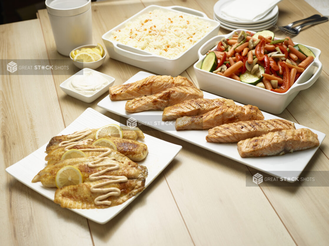 Catered assortment of fish, white rice and roasted vegetables on white servewear on a wooden background