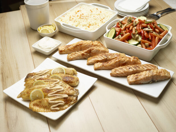 Catered assortment of fish, white rice and roasted vegetables on white servewear on a wooden background