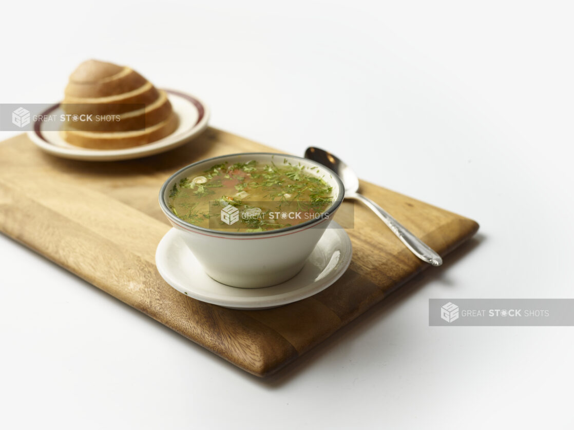 Bowl of matzo ball soup with plate of rye bread in the background on a wooden board on a white background