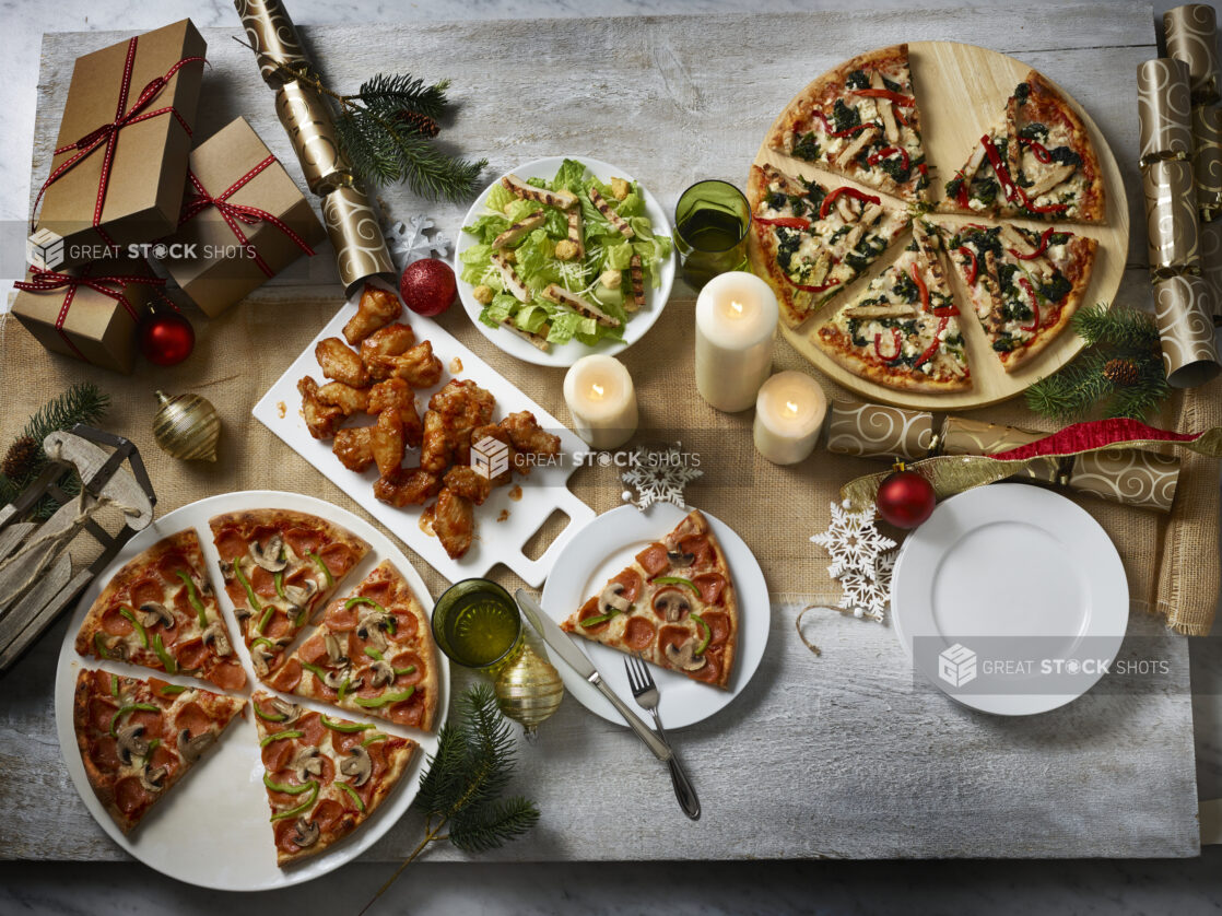 Holiday themed collection of pizzas, wings and a salad, overhead on a wooden table with a burlap runner