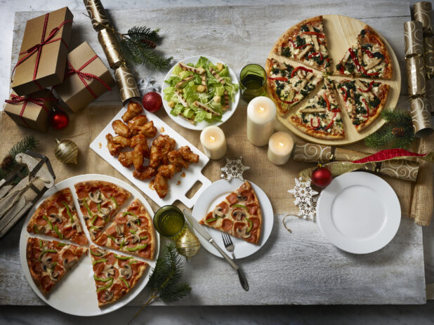 Holiday themed collection of pizzas, wings and a salad, overhead on a wooden table with a burlap runner