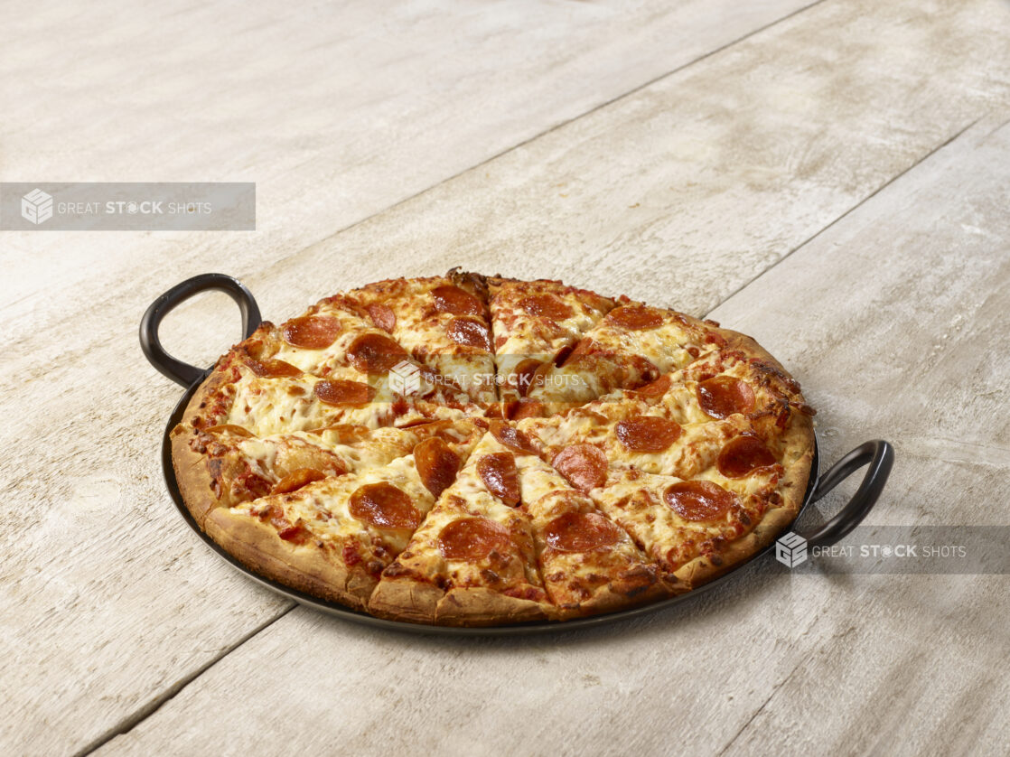 Sliced pepperoni pizza in a black cast iron pan on a wooden background