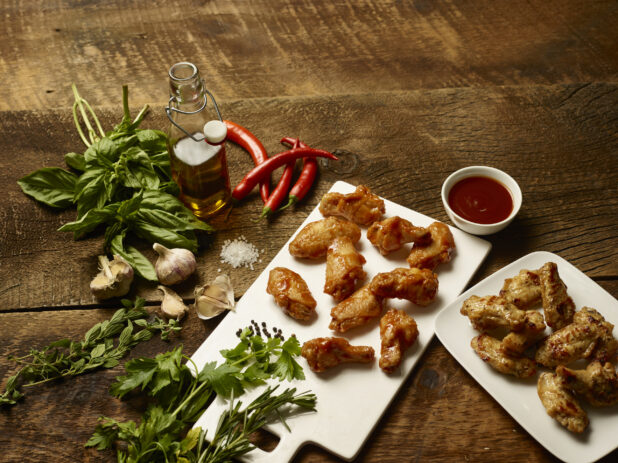 Board of sauced chicken wings with plate of plain wings overhead surrounded by fresh ingredients on a dark wooden background