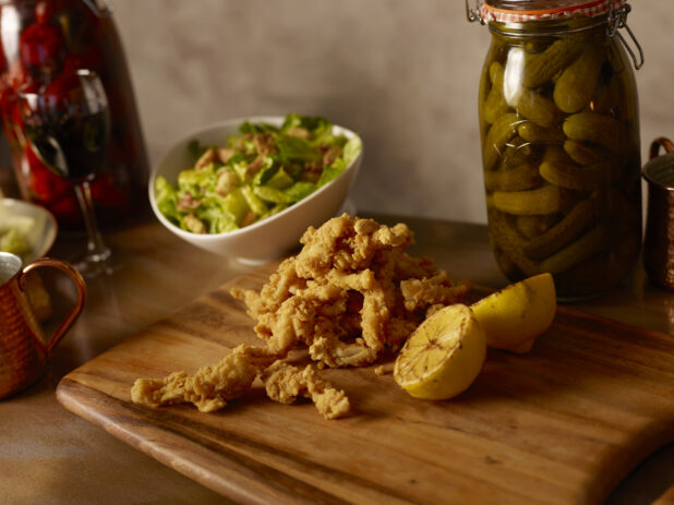 Fried calamari and grilled lemon on a wooden board with a bowl of salad behind