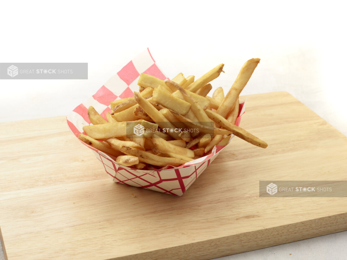 Basket of french fries in a lined red and white cardboard takeout container on a wooden cutting board