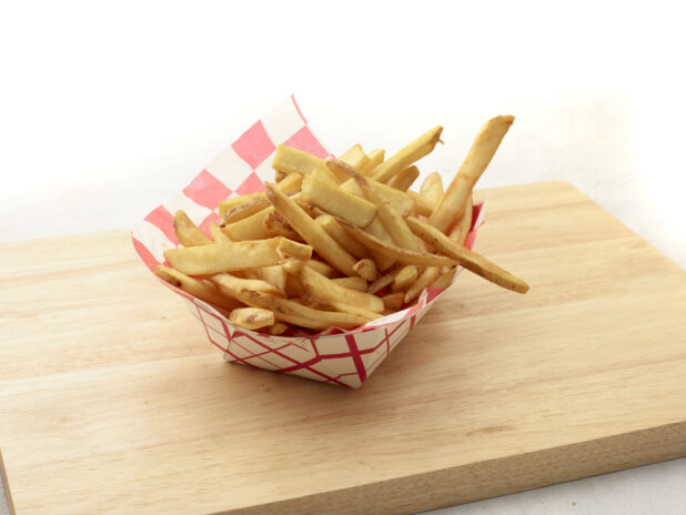 Basket of french fries in a lined red and white cardboard takeout container on a wooden cutting board