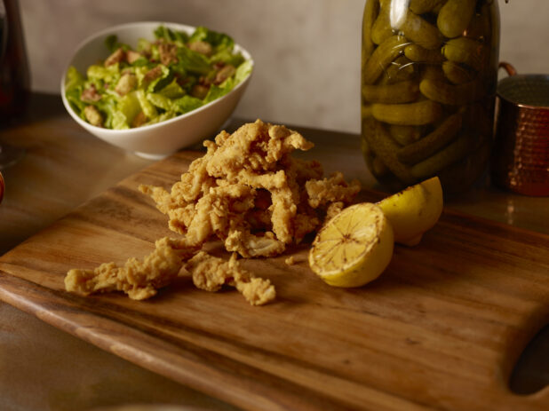 Fried calamari and grilled lemon on a wooden board with a bowl of salad in behind