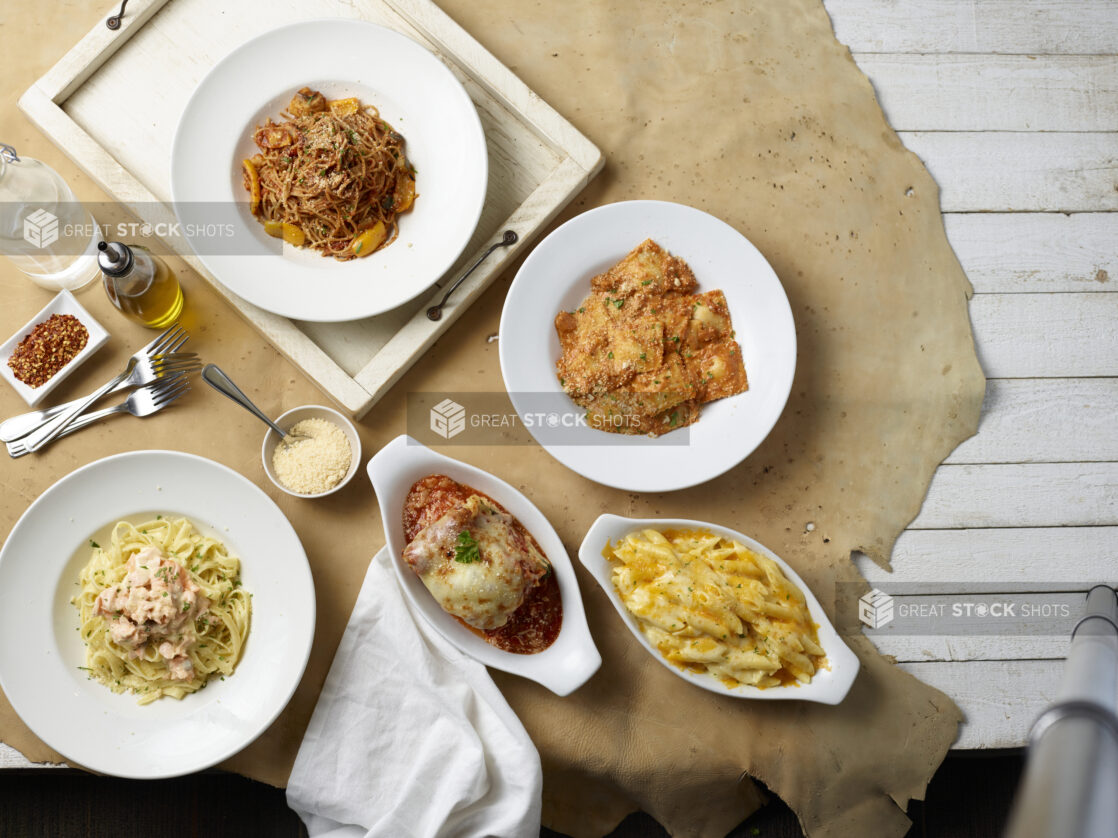 Overhead view of various pasta dishes with condiments on a cork background