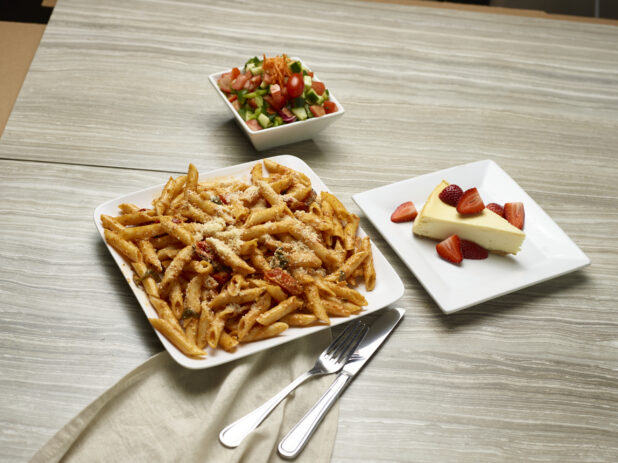 Penne in tomato sauce with a side salad and cheesecake for dessert on a wooden background