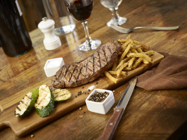 Grilled New York strip steak with side of grilled zucchini and frites on a wooden board with red wine