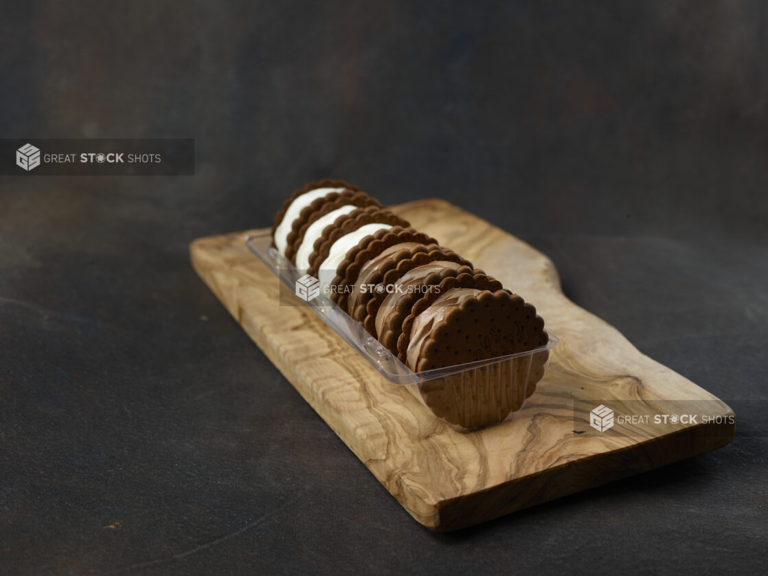 Vanilla and chocolate ice cream sandwiches in a plastic container on a wooden board on a dark background