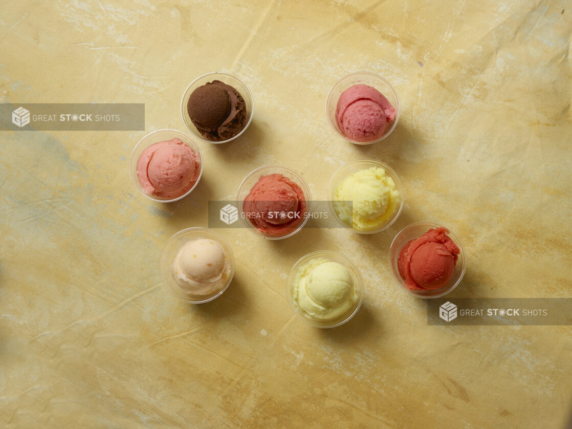 Various falvors of gelato / ice cream scooped into clear bowls, overhead, on a yellow background