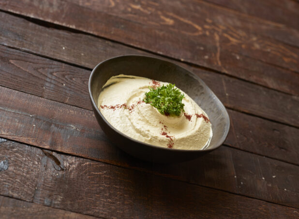 Bowl of hummus garnished with olive oil, paprika and fresh parsley in a brown bowl on a dark wooden background