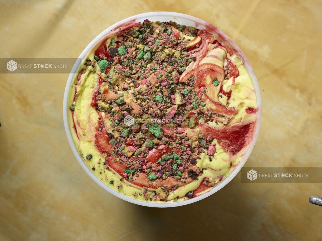 Overhead view of a large tub of rainbow ice cream with cookie / cake crumble and strawberry swirl on a yellow background