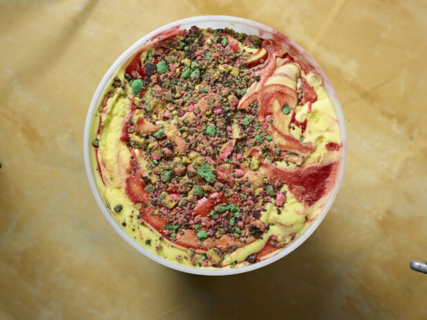 Overhead view of a large tub of rainbow ice cream with cookie / cake crumble and strawberry swirl on a yellow background