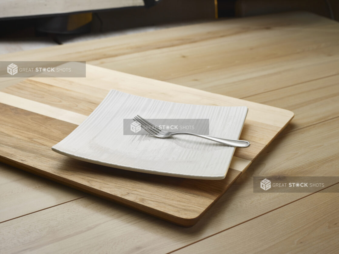 A fork on a square white plate sitting on a wooden cutting board