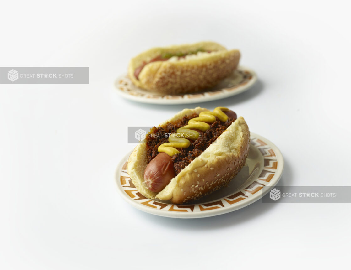 Two loaded hot dogs with mustard on sesame seed buns on diner plates, white background