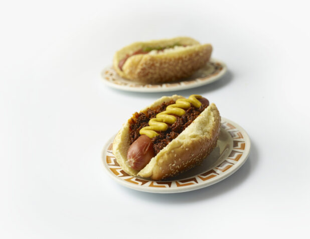 Two loaded hot dogs with mustard on sesame seed buns on diner plates, white background