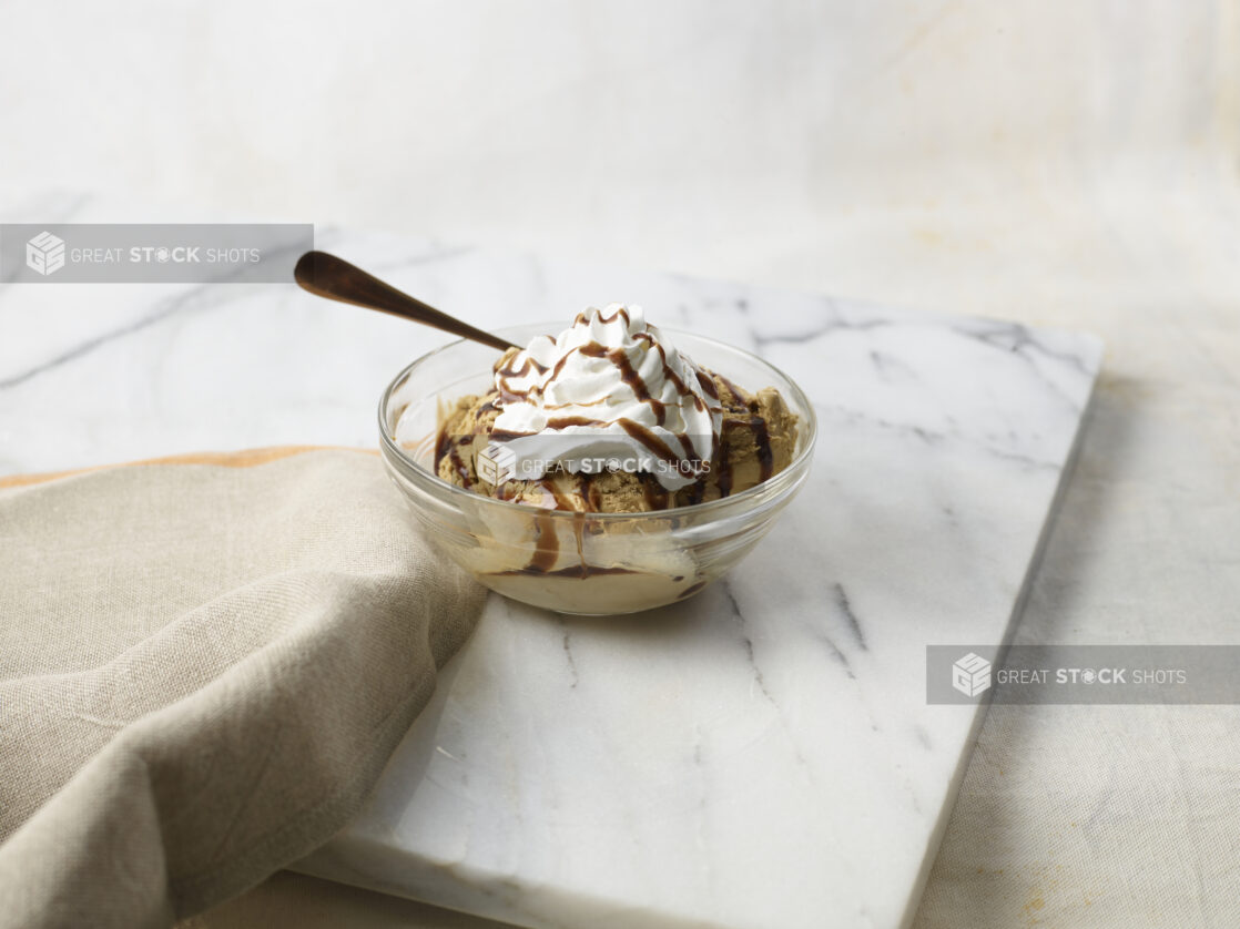 Glass bowl of ice cream with whipped cream on top, drizzled with chocolate sauce on a marble background