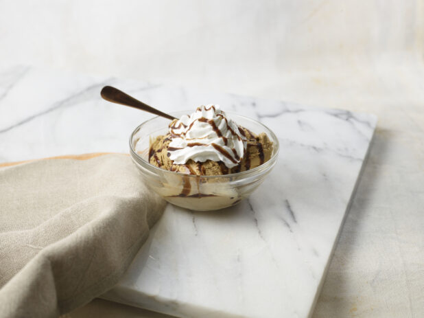 Glass bowl of ice cream with whipped cream on top, drizzled with chocolate sauce on a marble background