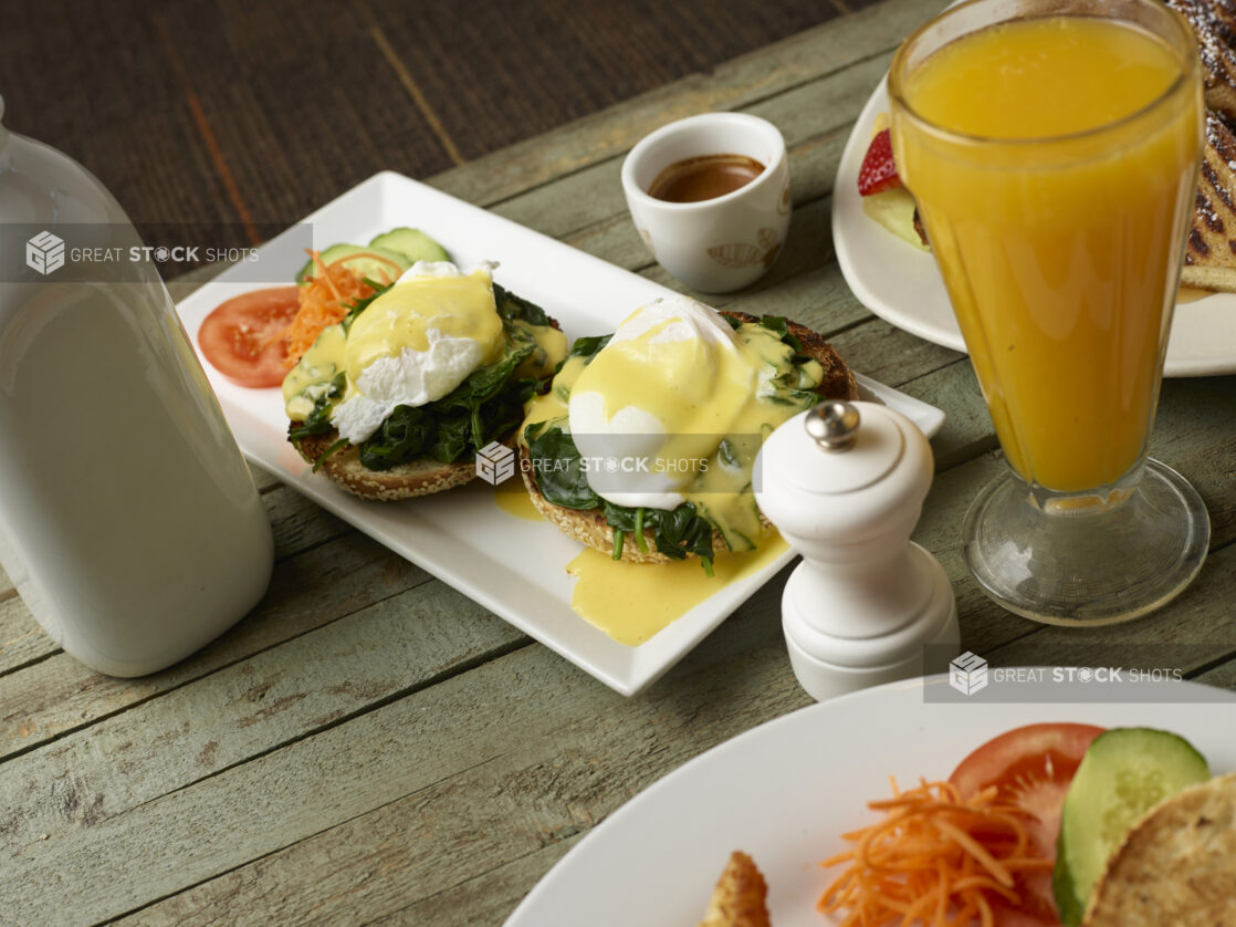 Eggs Benedict Florentine-style on a bagel on an oblong white plate with orange juice on a wooden background