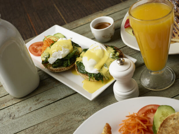 Eggs Benedict Florentine-style on a bagel on an oblong white plate with orange juice on a wooden background