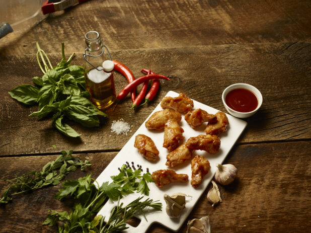 Board of sauced chicken wings overhead surrounded by fresh ingredients on a dark wooden background