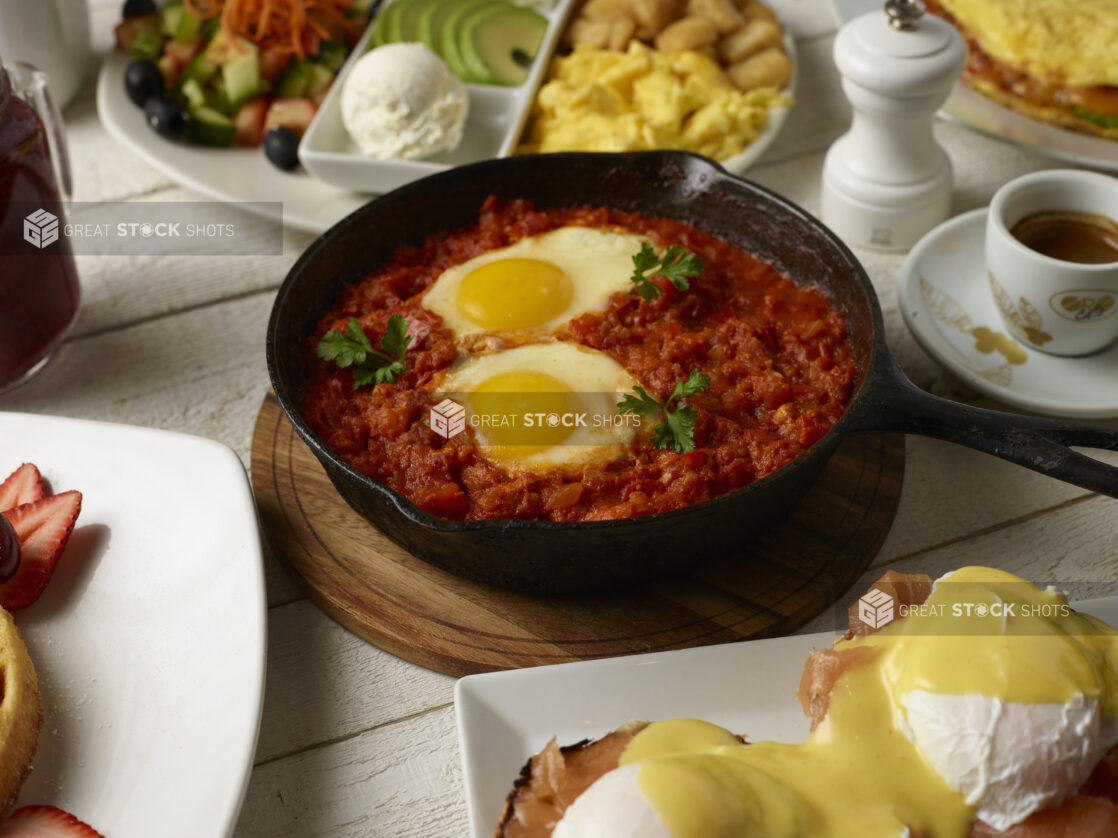 Various breakfast / brunch options around a skillet of shakshuka on a light wooden background