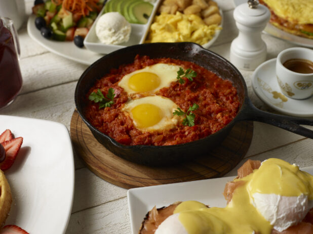 Various breakfast / brunch options around a skillet of shakshuka on a light wooden background