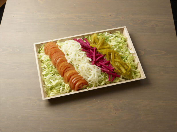 Wooden catering platter with tomatoes, onions, pickles and turnip on a bed of lettuce on top of a wooden background