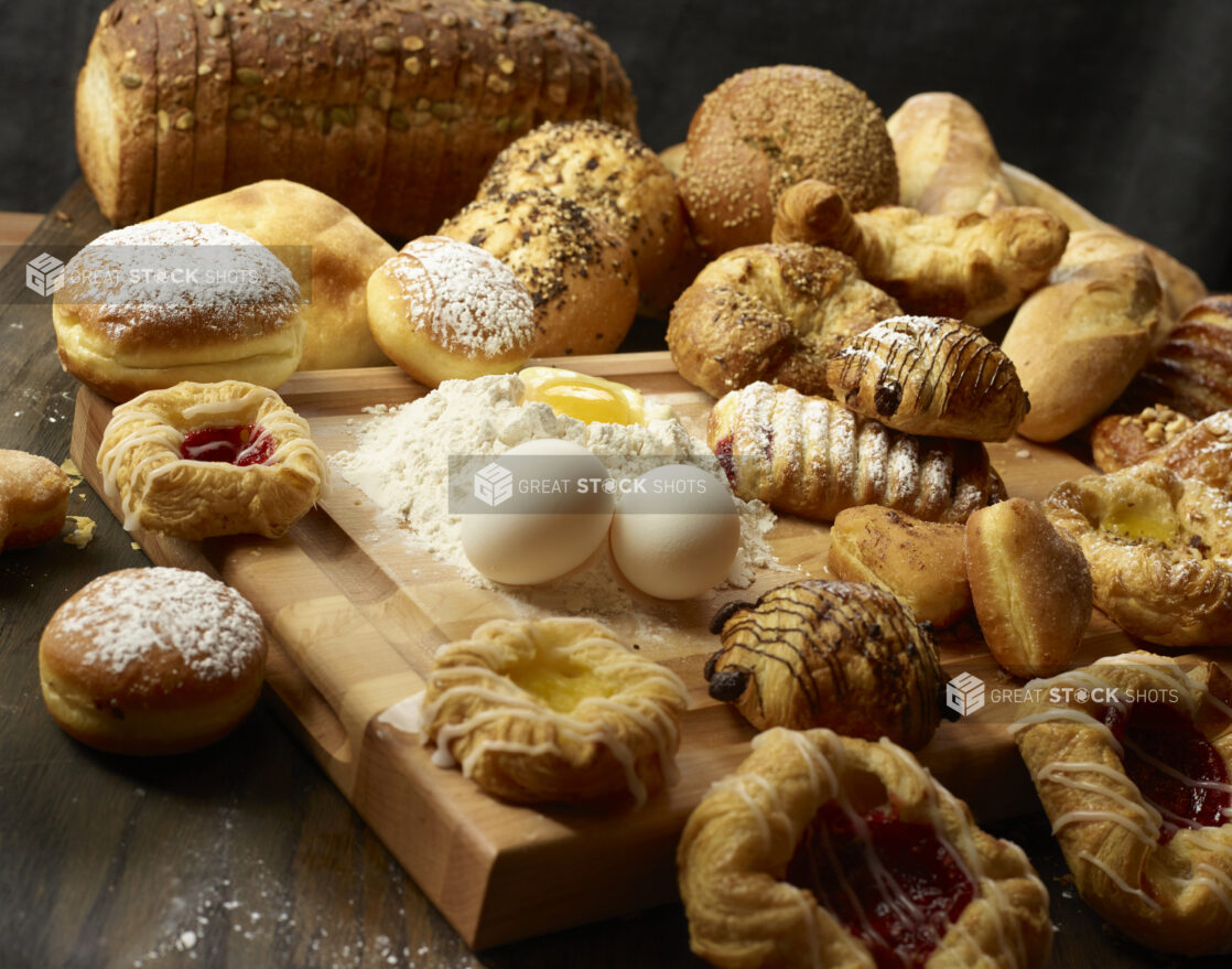 Collection of various breads and pastries with fresh eggs and flour with an egg well on a wooden block