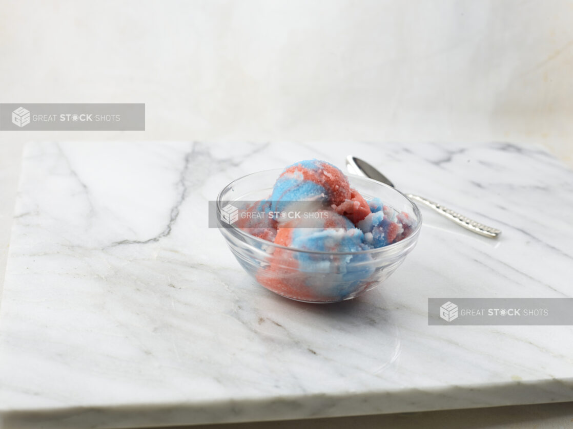 Multicolored gelato in a glass bowl with spoon on a marble background