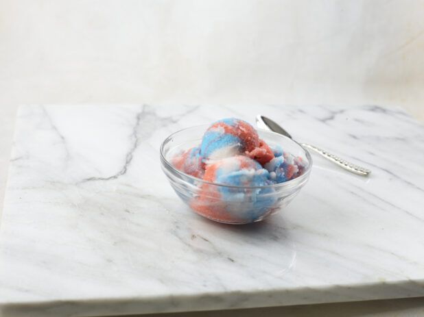 Multicolored gelato in a glass bowl with spoon on a marble background