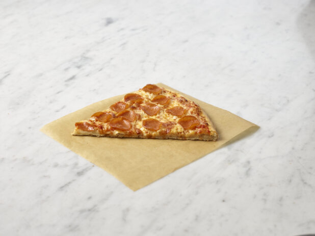 Slice of pepperoni pizza on parchment paper on top of a marble background