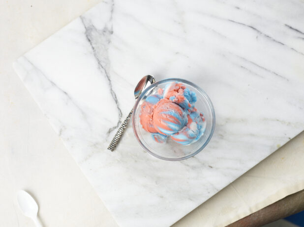 Multicolored gelato in a glass bowl with spoon on a marble background